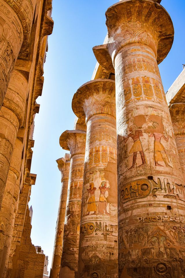 Columnas egipcias antiguas con jeroglíficos y tallas pintadas se elevan hacia un cielo azul brillante.