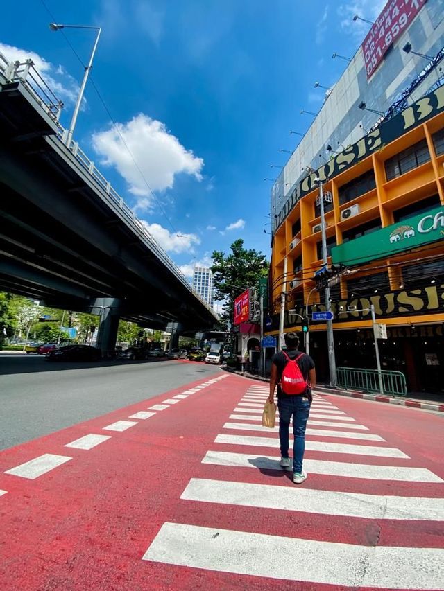 Una persona con una mochila roja camina por un paso de cebra rojo y blanco a lo largo de una calle de la ciudad junto a un paso elevado.