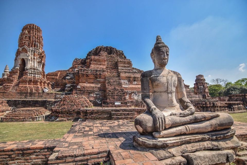 Una estatua de Buda de piedra se sienta en pose meditativa frente a antiguas ruinas de templo de ladrillo rojo bajo un cielo azul claro.