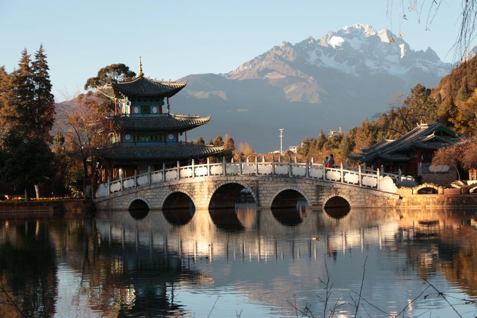 Una pagoda tradizionale e un ponte ad arco in pietra si riflettono nell'acqua calma, con una montagna innevata sullo sfondo.