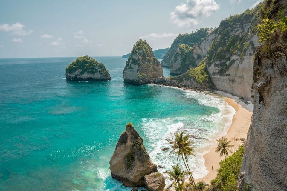 Vista elevada de una cala tropical con playa de arena, agua turquesa y grandes formaciones rocosas bajo un cielo azul.