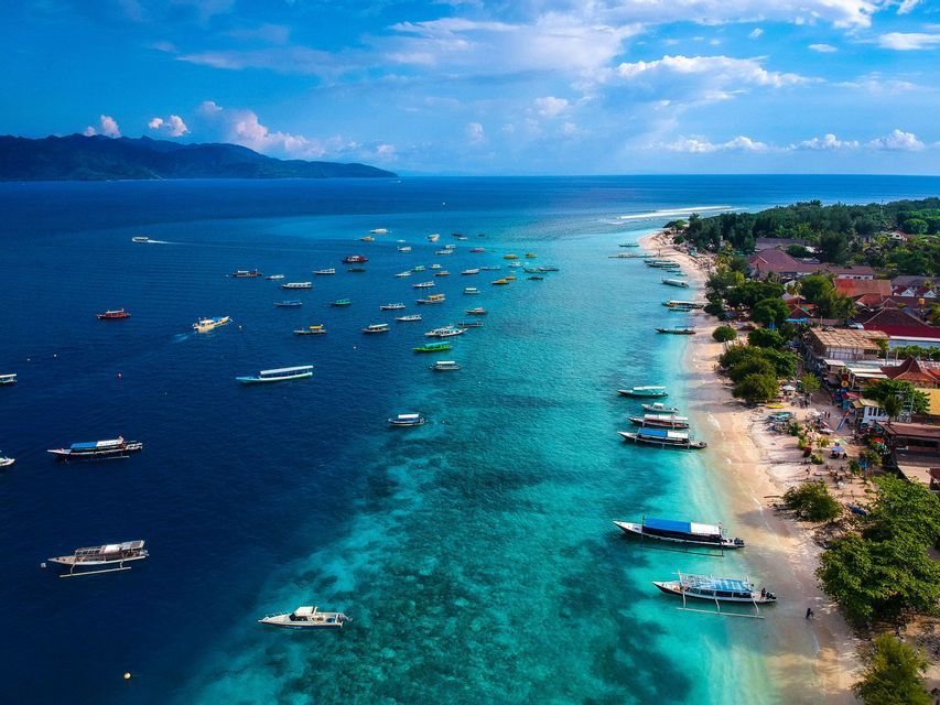 Una vista aérea muestra decenas de barcos flotando en agua cristalina color turquesa junto a una playa de arena bordeada de árboles y edificios.