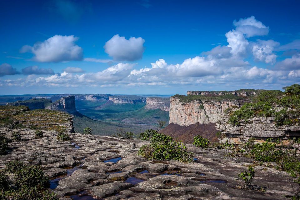 A rocky plateau with small puddles of water overlooks a vast green valley with mesas under a blue sky with white clouds.