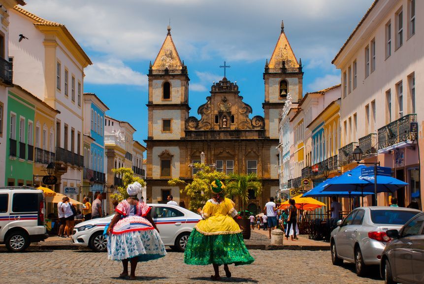 Two women in colorful traditional dresses stand on a cobblestone street in front of a historic church with twin bell towers.
