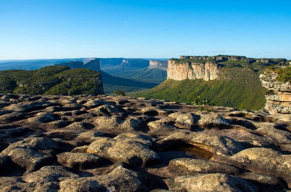 A view from a rocky plateau overlooking a deep green valley with large, flat-topped mountains in the distance under a clear blue sky.