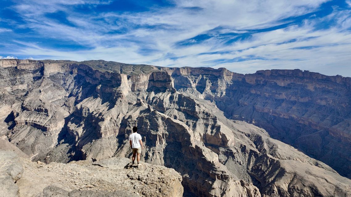 Eine Person steht am Rande einer Klippe und blickt über eine tiefe, felsige Schlucht unter blauem Himmel mit Wolken.