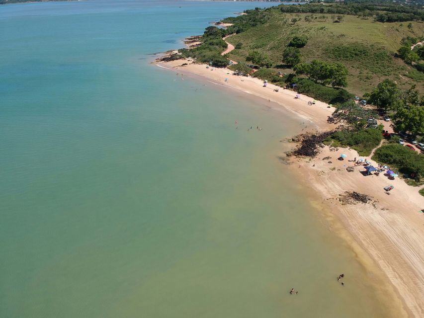 Una vista aérea de una playa de arena que se curva a lo largo de una costa con agua turquesa y colinas verdes. La gente está nadando y relajándose en la orilla.