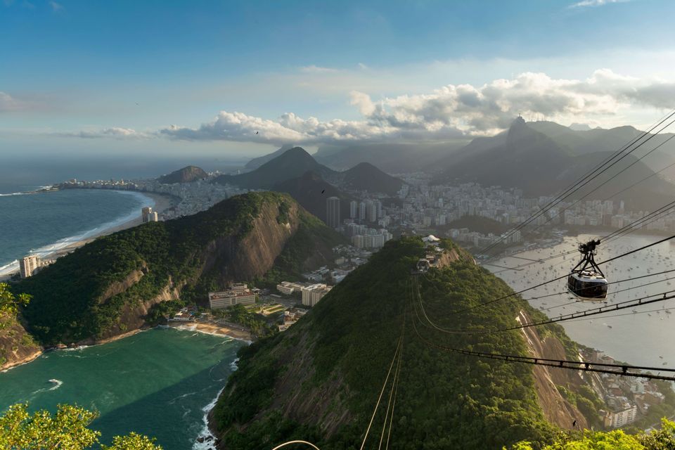Un teleférico viaja sobre montañas verdes, ofreciendo una vista panorámica de una ciudad costera, una bahía con barcos y el océano.