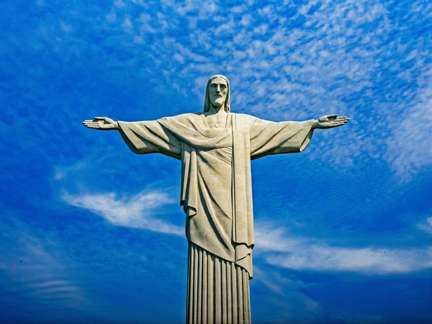La estatua del Cristo Redentor con los brazos extendidos, vista desde un ángulo bajo contra un cielo azul brillante con nubes dispersas.