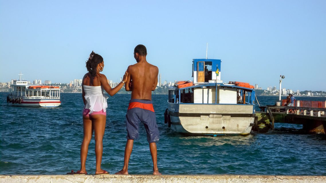 A man and a woman stand on a seawall looking out at boats on the water, with a city skyline visible in the distance.