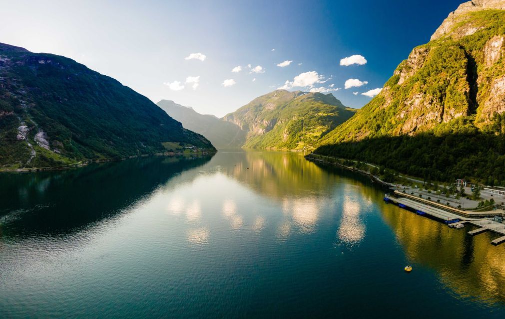 Weitwinkelaufnahme eines Fjords mit ruhigem, spiegelndem Wasser, umgeben von steilen, grünen Bergen unter blauem Himmel mit einigen Wolken.