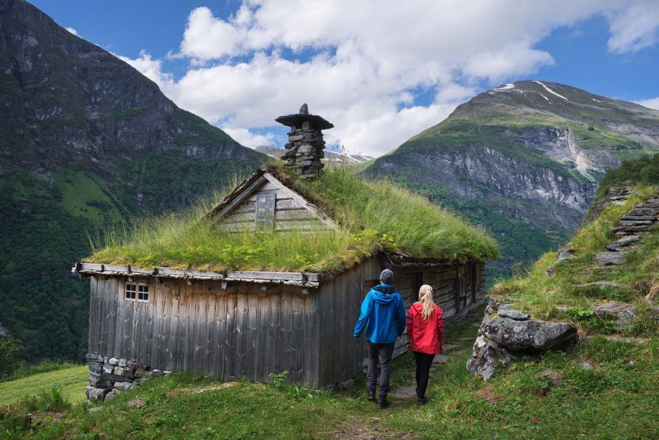 Ein Paar spaziert auf einem Pfad zu einer Holzhütte mit Grasdach, umgeben von steilen grünen Bergen unter blauem Himmel.