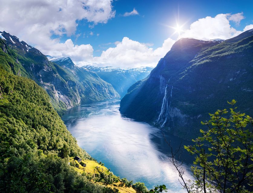 Ein breiter Fjord schlängelt sich durch steile, bewaldete Berge, mit einem Wasserfall auf der einen Seite und der Sonne, die darüber scheint.