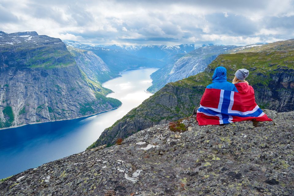 Zwei Personen, eingehüllt in eine norwegische Flagge, sitzen auf einer felsigen Klippe mit Blick auf einen weiten Fjord und ferne schneebedeckte Berge.