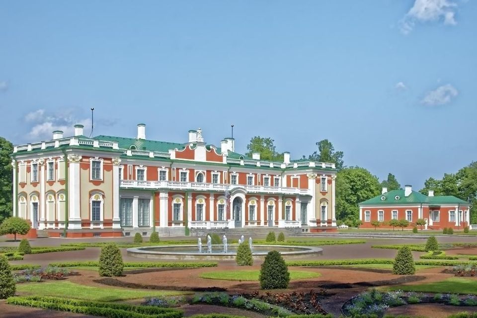 Ein großes, rot-cremefarbenes Barockschloss mit grünem Dach, gesehen über einen formalen Garten mit einem runden Brunnen und Hecken.