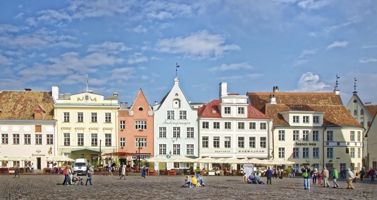 Ein Panoramablick auf einen europäischen Kopfsteinpflasterplatz mit farbenfrohen, historischen Gebäuden und Menschen, die einen sonnigen Tag genießen.