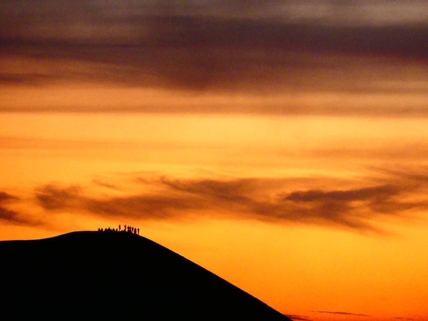 Un groupe WeRoad se profile en silhouette au sommet d'une grande dune de sable, sur fond de spectaculaire coucher de soleil orangé.