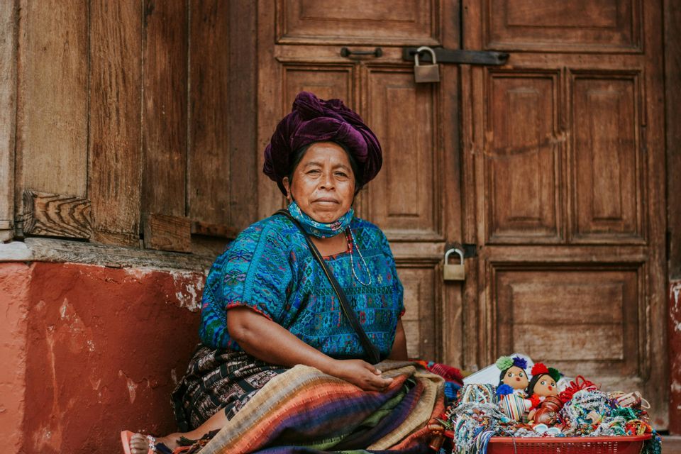 Une femme en tenue traditionnelle colorée et coiffe violette est assise devant une porte en bois avec un panier de poupées faites à la main.