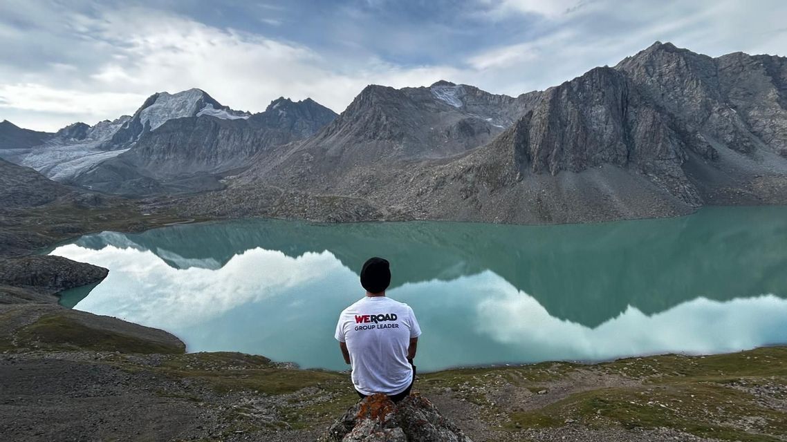 From behind, a WeRoad Group Leader sits on a rock overlooking a turquoise alpine lake that reflects snow-capped mountains.