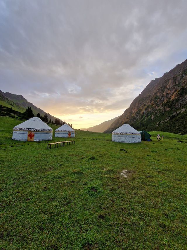 Drei traditionelle Jurten stehen auf einer grünen Wiese in einem Bergtal bei bewölktem Sonnenuntergang.