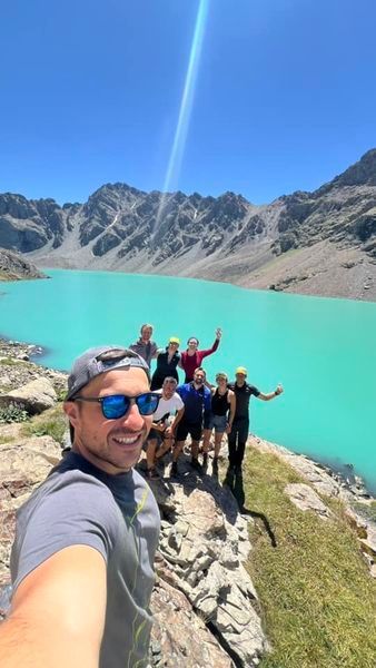 Un gruppo WeRoad si scatta un selfie su uno sperone roccioso che domina un lago alpino turchese e montagne impervie.