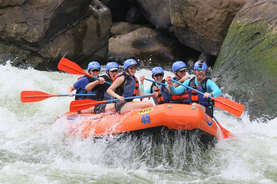Eine WeRoad-Gruppe lacht und paddelt in Helmen und Schwimmwesten mit einem orangen Schlauchboot durch Wildwasser-Stromschnellen.