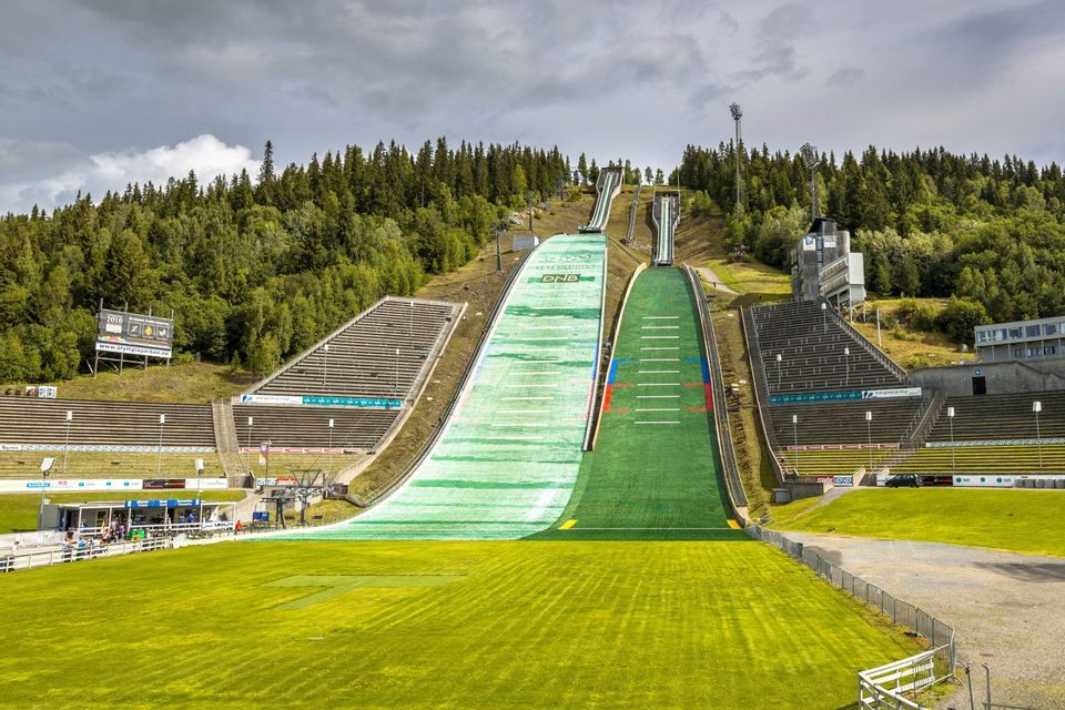 Zwei große Skisprungschanzen mit grünen Kunstrasen-Landebahnen stehen in einem leeren Stadion, umgeben von einem dichten Wald unter einem bewölkten Himmel.