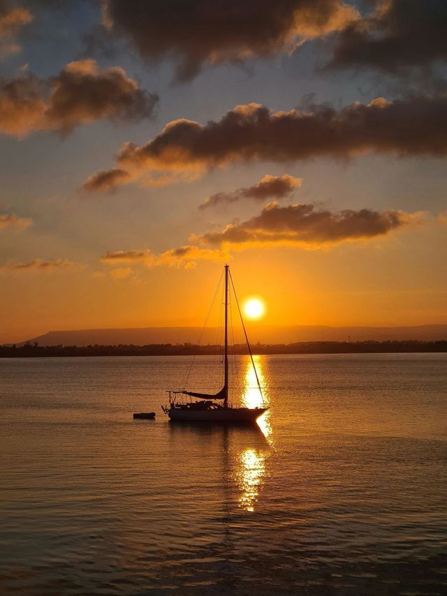 Una barca a vela galleggia sull'acqua calma mentre il sole tramonta, proiettando un riflesso dorato sulla superficie.