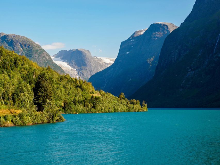 Ein leuchtend blauer Fjord mit grünen, bewaldeten Ufern liegt am Fuße großer, schneebedeckter Berge unter einem klaren Himmel.