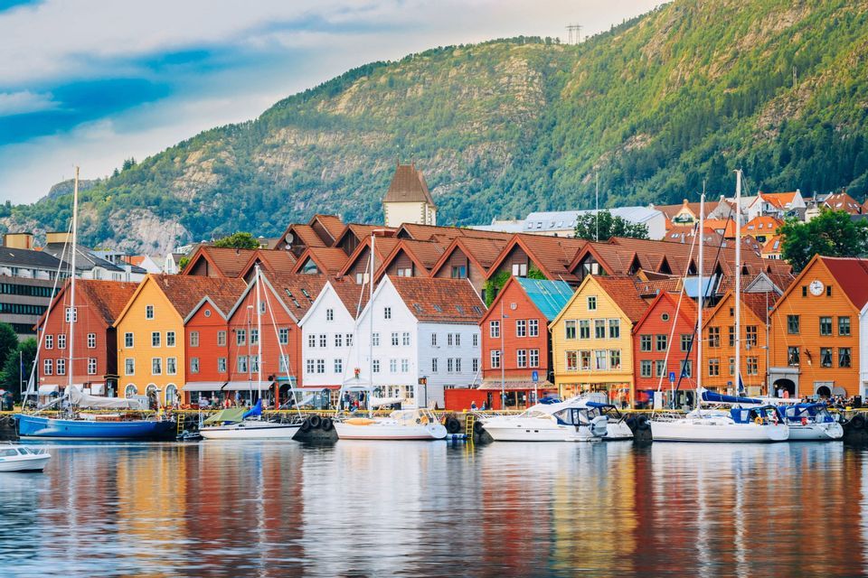 Bunte Holzhäuser und Boote säumen das Ufer, mit Spiegelungen im Wasser und einem grünen Hügel im Hintergrund.