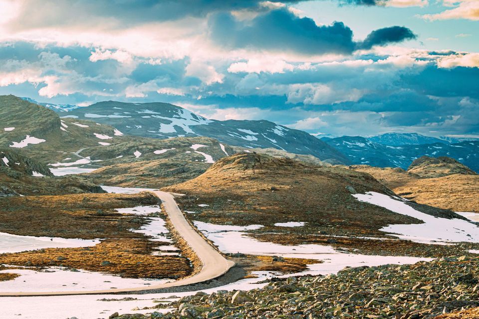 Eine schmale Straße schlängelt sich durch eine felsige, bergige Landschaft mit Schneeflecken unter einem bewölkten Himmel.