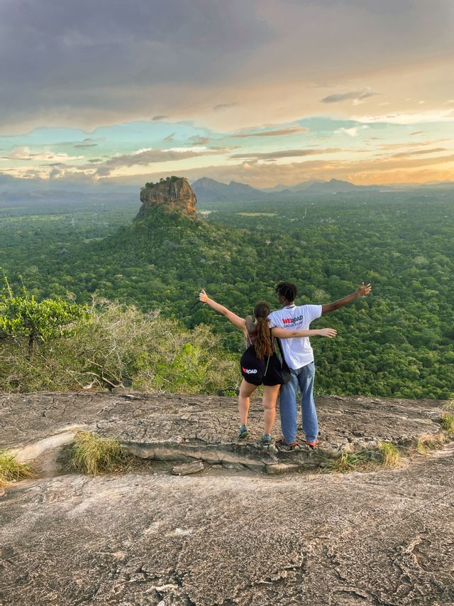 Zwei Personen einer WeRoad-Gruppenreise umarmen sich mit offenen Armen auf einer Klippe, mit Blick auf einen weiten Wald und eine ferne Felsenfestung.