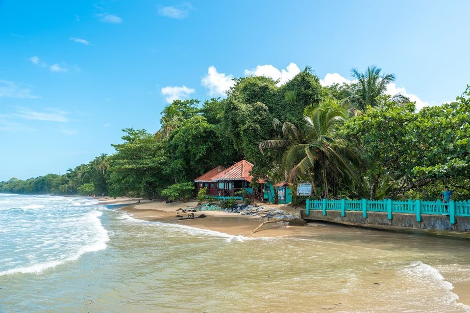 Una casa con il tetto rosso si trova su una spiaggia sabbiosa accanto a un molo turchese, con una lussureggiante foresta tropicale sullo sfondo.