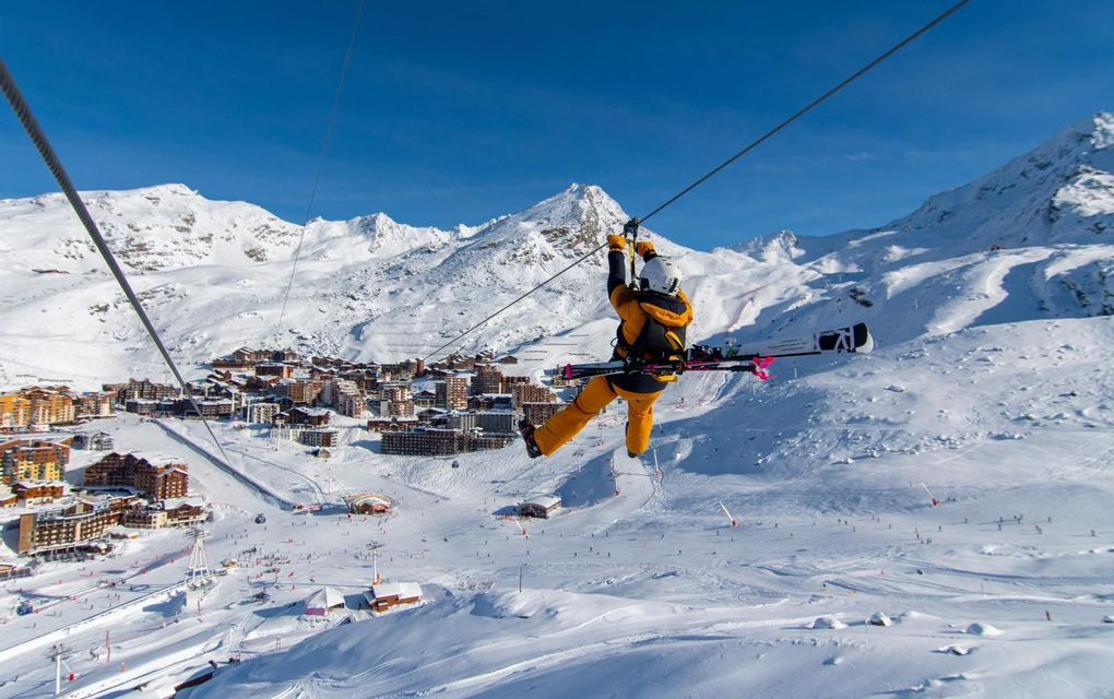 A person wearing ski gear glides down a zip line with skis on their back, high above a snowy mountain village.