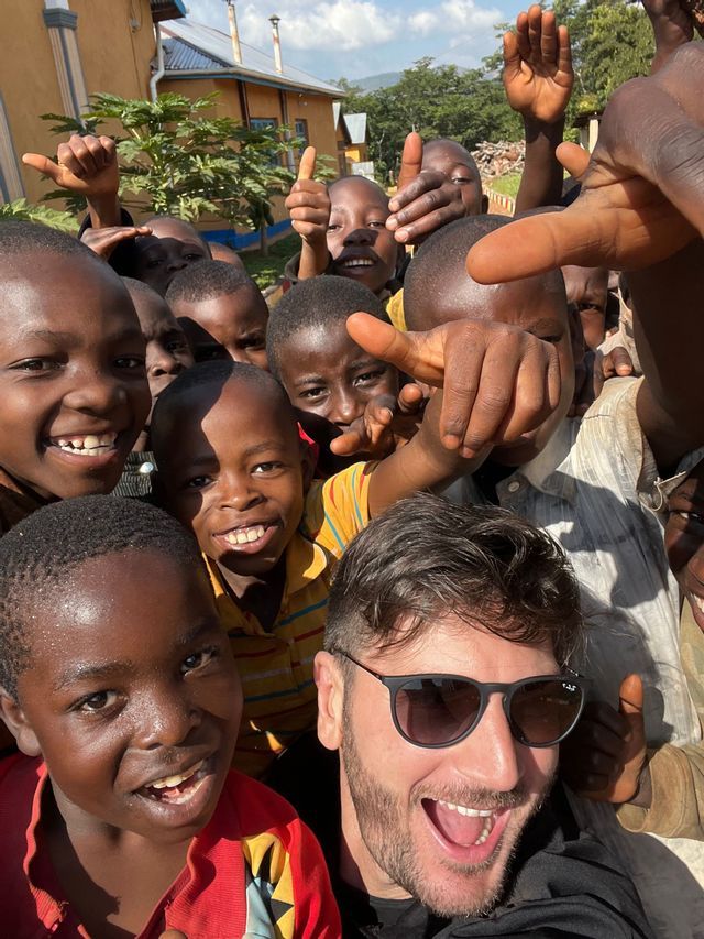 Un uomo sorridente in viaggio con WeRoad si scatta un selfie con un folto gruppo di bambini che si sono radunati intorno, sorridendo e in posa.