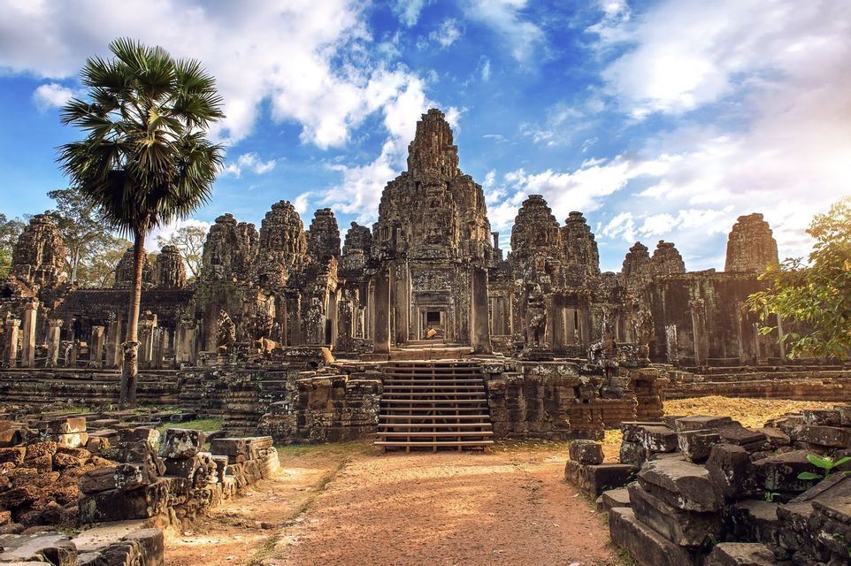 A large, ancient stone temple with carved towers and a central staircase, with a palm tree on the left under a partly cloudy sky.
