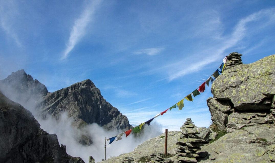 Bandiere di preghiera colorate sono tese tra tumuli di pietre su una cima di montagna, con nuvole che vorticano nella valle sottostante.