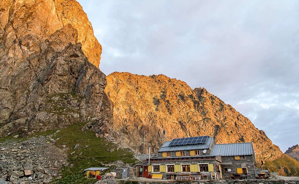 Un rifugio di montagna in pietra con pannelli solari sul tetto, adagiato su una montagna rocciosa illuminata dal sole dorato.