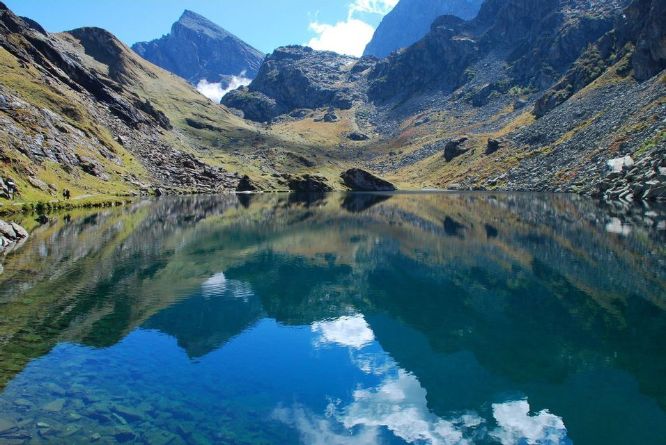 Un limpido lago alpino riflette perfettamente le montagne rocciose circostanti, con un viaggio di gruppo WeRoad che cammina lungo la sua riva.