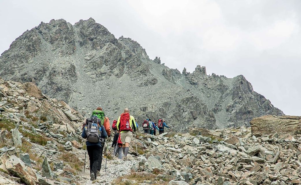 Un viaggio di gruppo WeRoad con zaini e bastoni da trekking risale un sentiero di montagna roccioso verso una cima frastagliata sotto un cielo coperto.