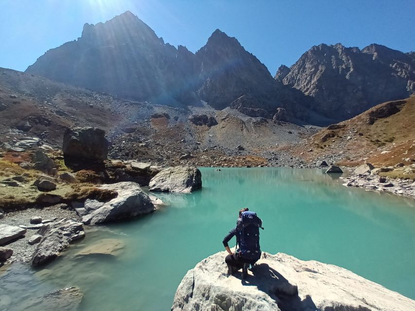 Un escursionista con un grande zaino si accovaccia su una roccia, affacciandosi su un lago alpino turchese ai piedi di montagne frastagliate.