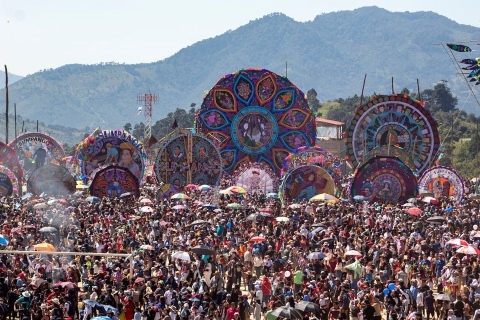 Una grande folla si raduna per un festival con aquiloni giganti, colorati e circolari, con montagne verdi sullo sfondo.