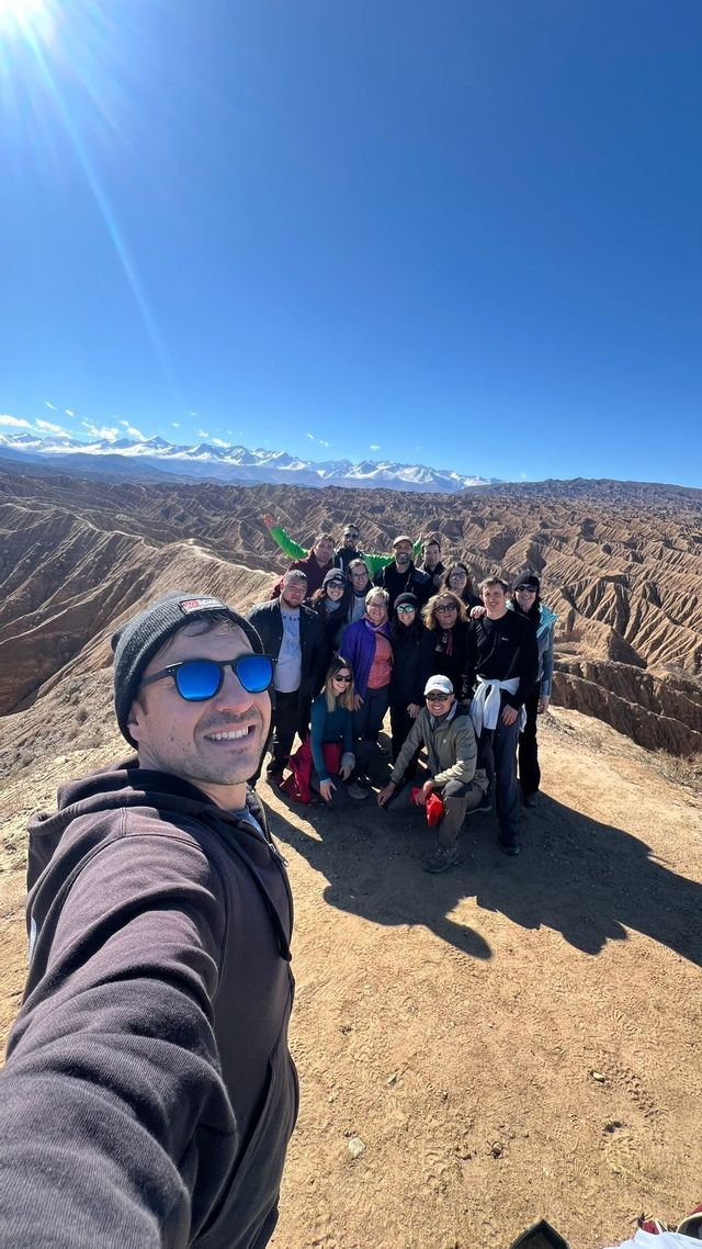 Un uomo si scatta un selfie con un gruppo WeRoad su una cresta che domina un canyon, con montagne innevate sotto un cielo soleggiato.