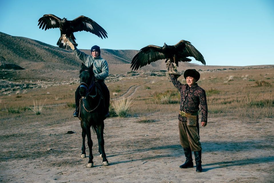 Ein Mann auf einem dunklen Pferd und ein stehender Mann halten zwei Adler mit ausgebreiteten Flügeln in einer kargen, hügeligen Landschaft.