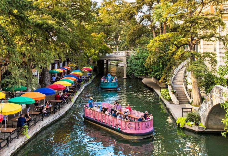 Barche turistiche colorate navigano lungo un canale fiancheggiato da alberi lussureggianti e caffè sul lungofiume con ombrelloni variopinti.