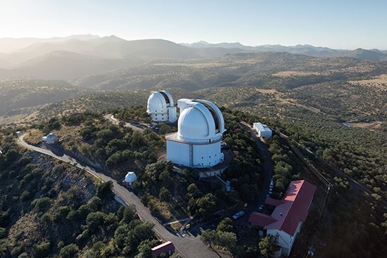 Veduta aerea di osservatori astronomici bianchi su una cima montuosa ricoperta di vegetazione, che domina un vasto paesaggio collinare.