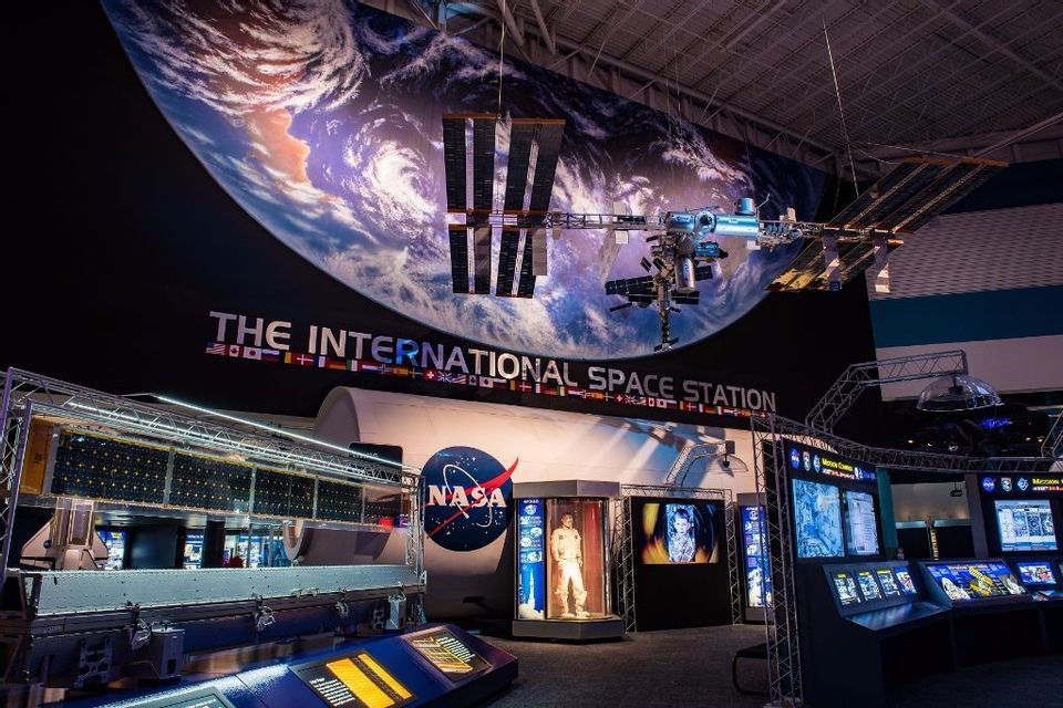 Una mostra interna della Stazione Spaziale Internazionale, con un grande modello appeso sopra un modulo NASA e display interattivi.