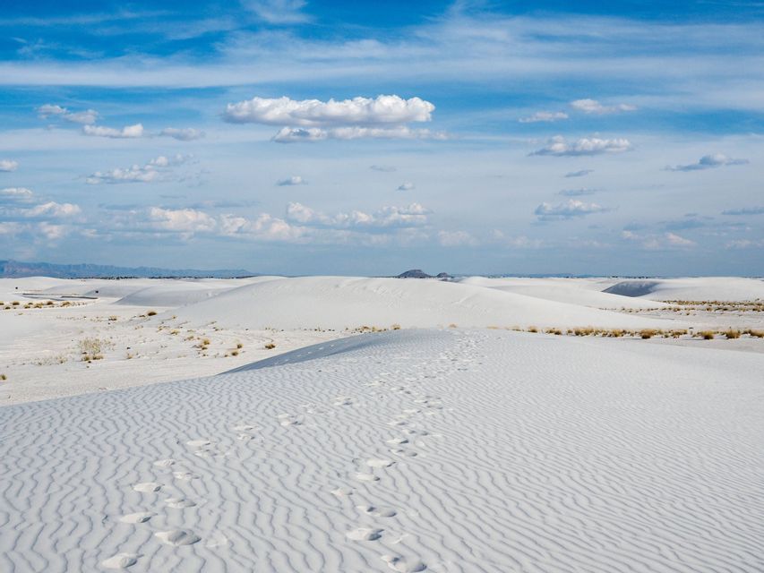 Un sentiero di impronte attraversa vaste dune di sabbia bianca ondulate, sotto un cielo azzurro e luminoso con nuvole bianche.