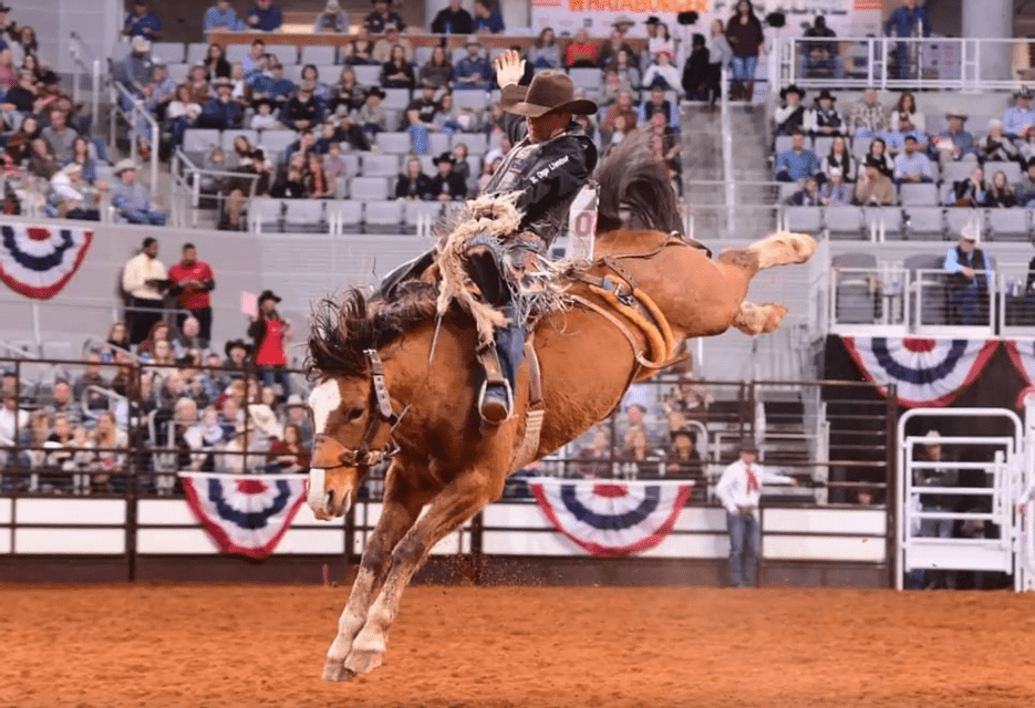 Un cowboy con un cappello alza una mano mentre cavalca un cavallo imbizzarrito in un'arena da rodeo sterrata, di fronte a una folla.