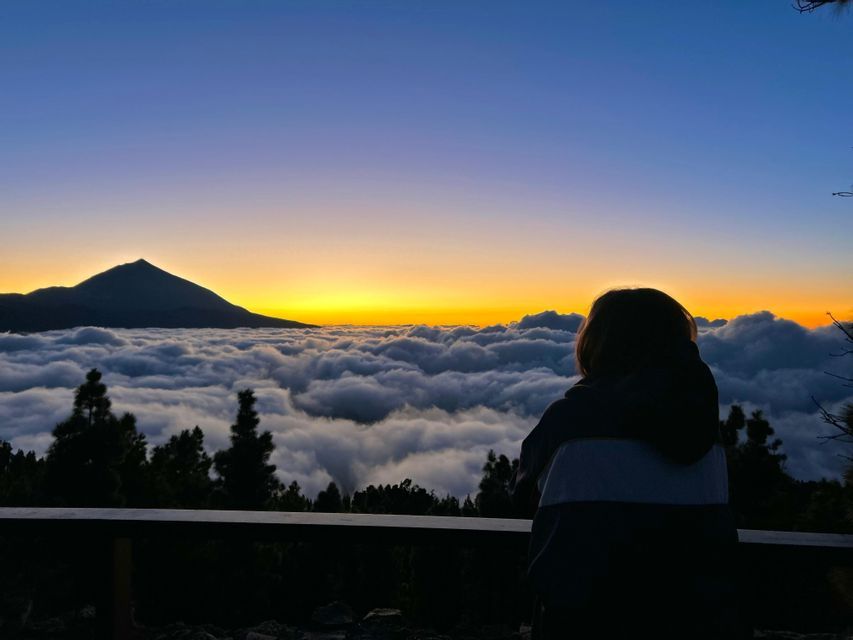 Una persona vista di spalle osserva una cima montuosa che si erge sopra un mare di nuvole al tramonto.